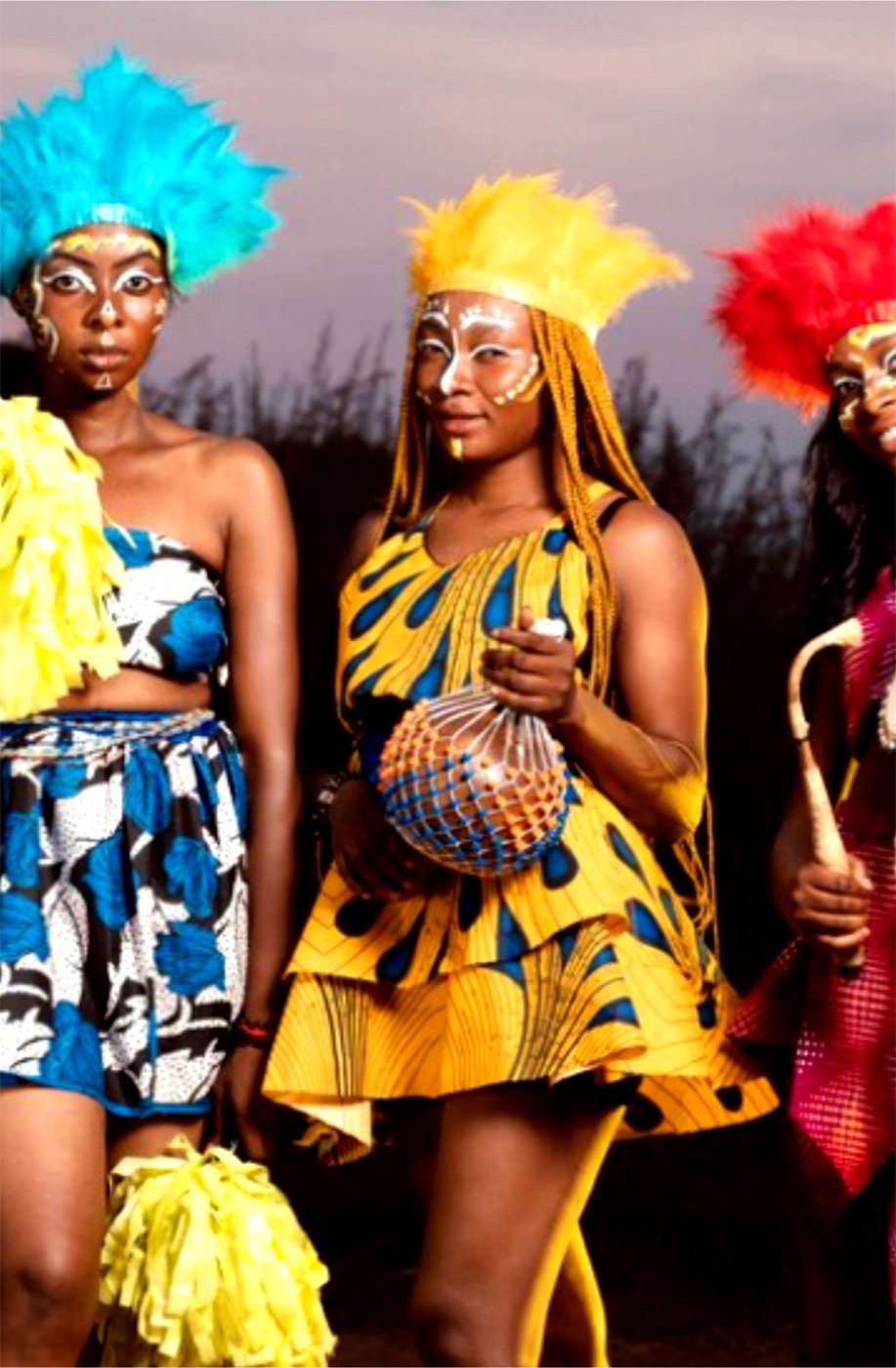 Three performers in feathered headdresses and African wax-print outfits holding a shekere and a drum